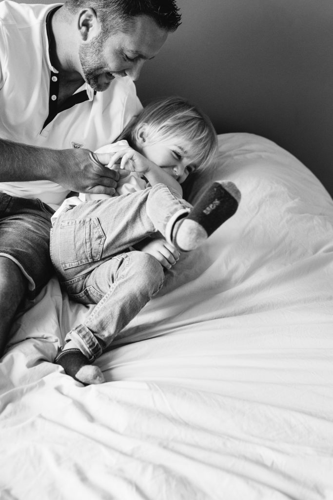 Photographie en noir et blanc d'un papa qui fait des chatouilles à son fils sur un lit